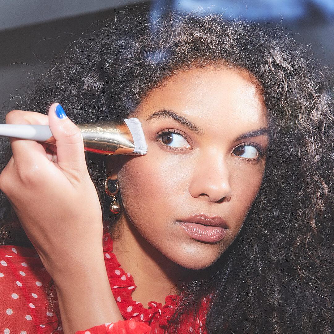 Beautiful model in a red and white polka dot blouse applying face makeup using a COMPLEX CULTURE makeup brush Mobile