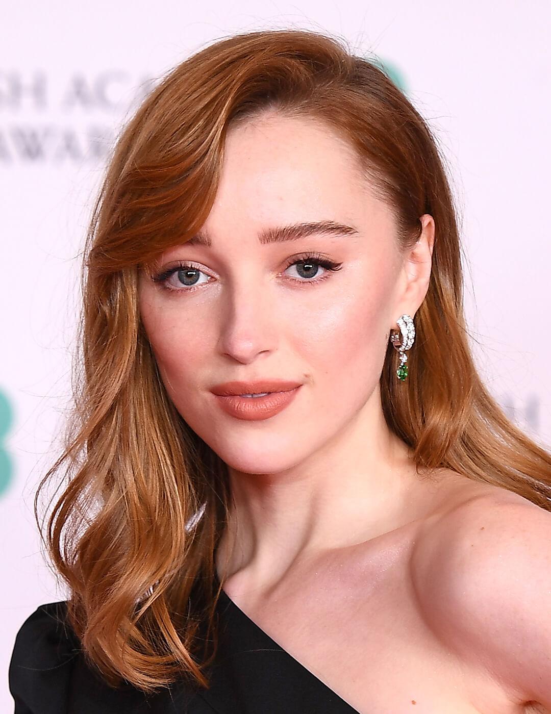 A photo of Phoebe Dynevor with a copper colored hair wearing a black dress Mobile