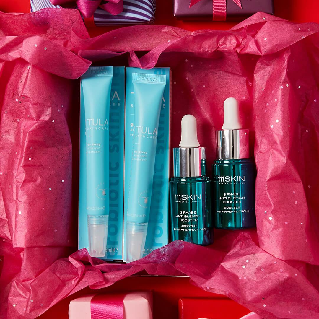 Flat lay image of two tubes of TULA SKINCARE Go Away Acne Spot Treatment and two bottles of 111SKIN 3 Phase Anti Blemish Booster inside a pink gift box on red background Desktop