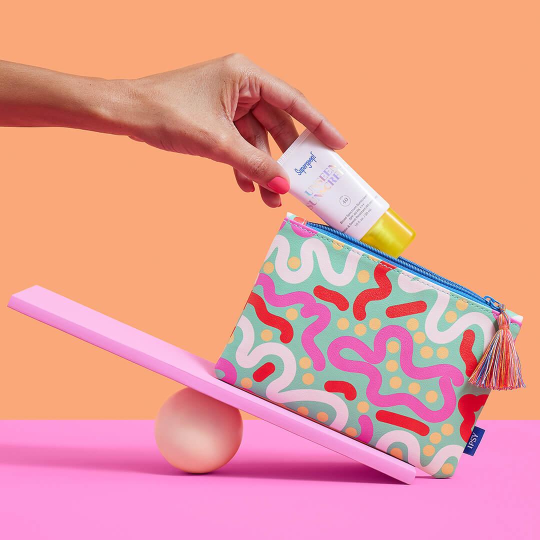 Woman's hand putting SUPERGOOP! inside a colorful makeup bag on a mini pink seesaw against orange and pink background Mobile