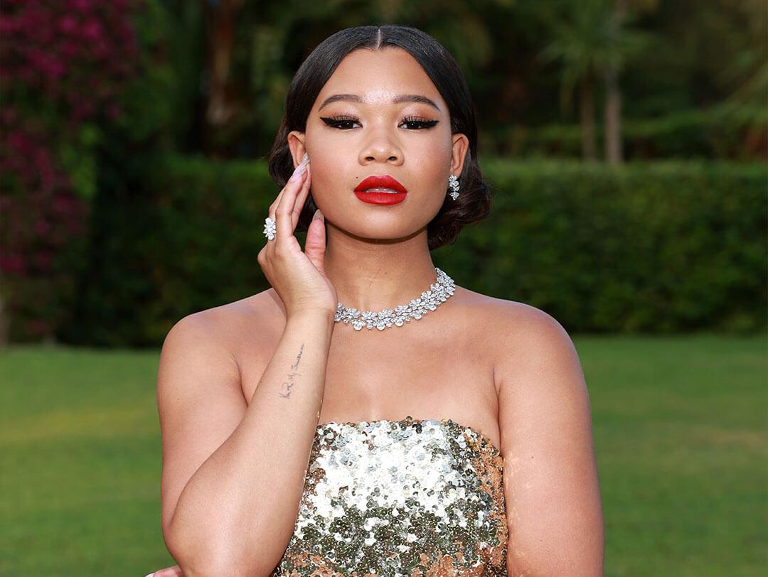 An image of Storm Reid, looking stunning in a gold sequined sleeveless dress, thick black eyeliner, red lipstick, and a set of earrings, rings, and necklaces, with her hair split in the center Desktop