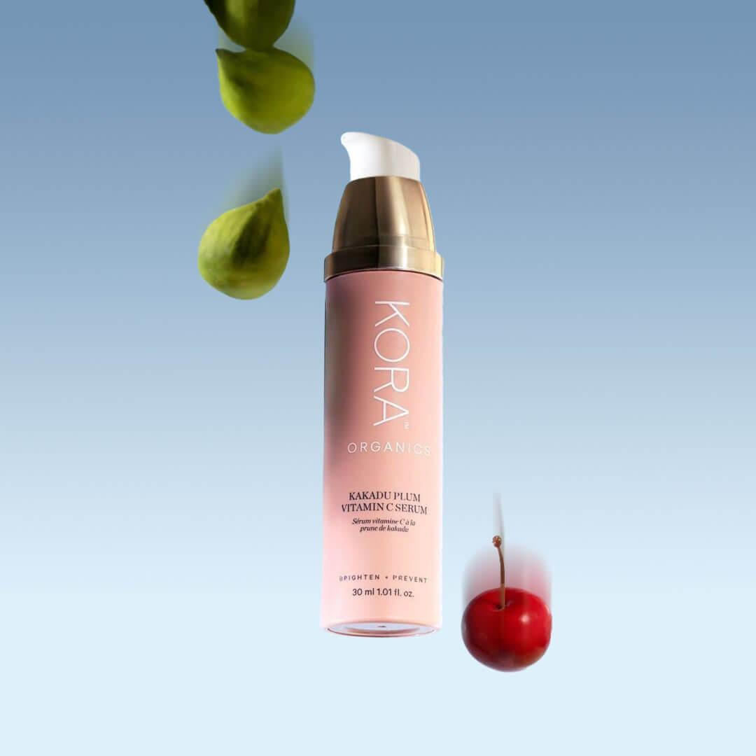 A pink and gold bottle of KORA ORGANICS Kakadu Plum Vitamin C Serum stands against a serene blue backdrop, with a fresh green leaf and a bright red fruit Desktop