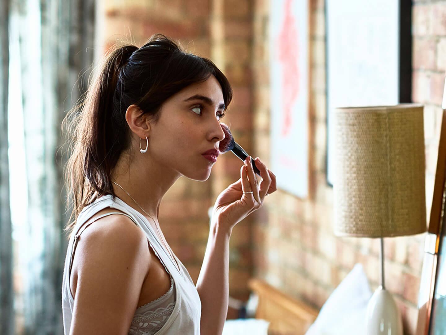 An image of a woman applying makeup with a brush on her face while holding a toast in front of a mirror Desktop