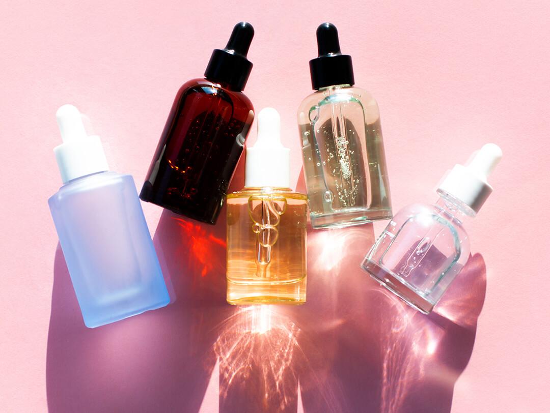 Row of bottles of different shapes, sizes, and colors with beauty products inside against pink background Desktop
