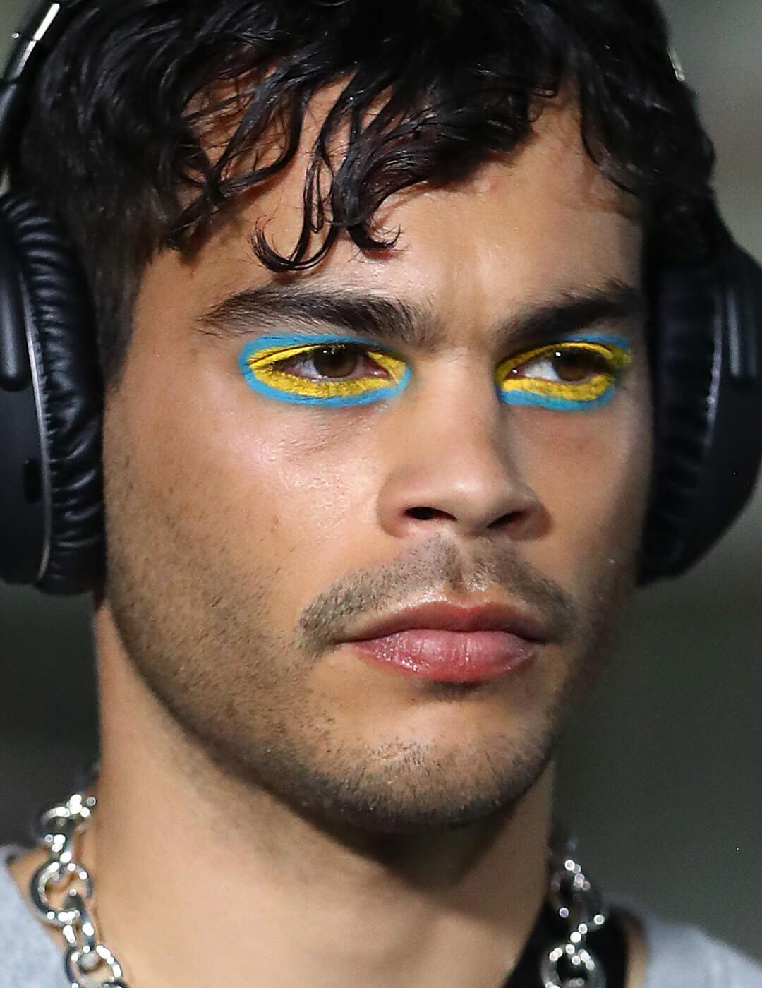 Male model wearing black headphones, silver necklace, and blue and yellow eyeshadow makeup Mobile