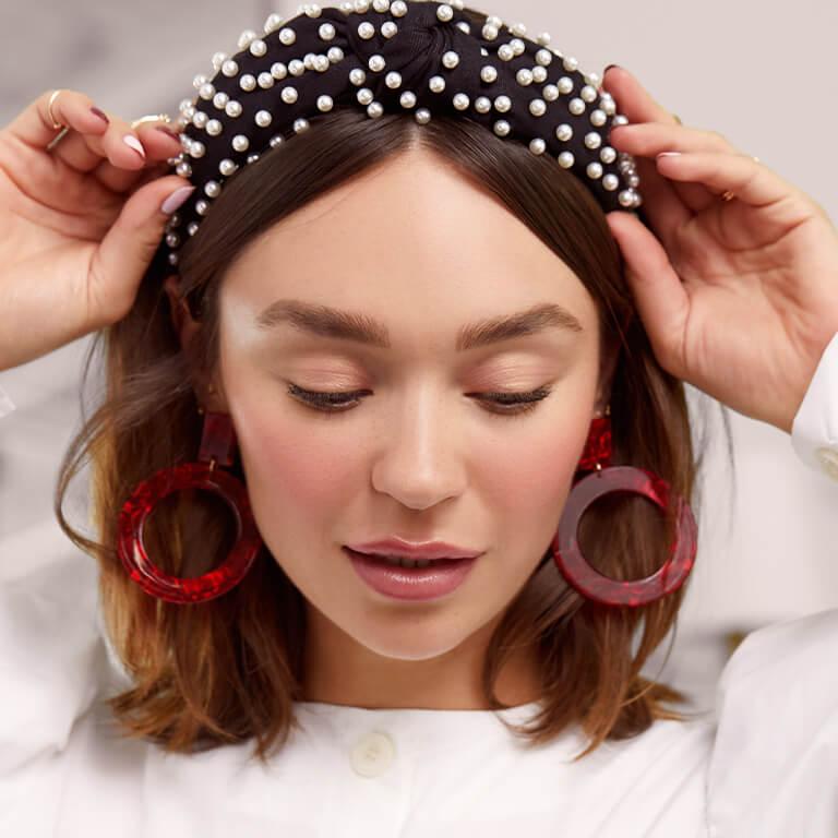 An image of a model with a no-makeup makeup look wearing bold red earrings while touching her pearl-studded headband Desktop