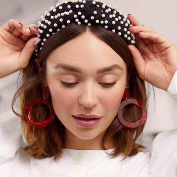 An image of a model with a no-makeup makeup look wearing bold red earrings while touching her pearl-studded headband Mobile