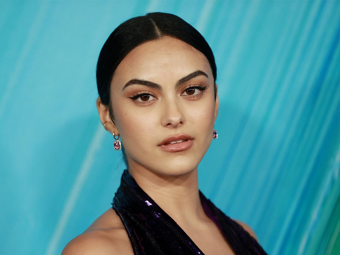 An image of Camila Mendes with her sleek blue-black hair, gazing at the camera, wearing black eyeliner and a stylish dark halter top purple dress Desktop