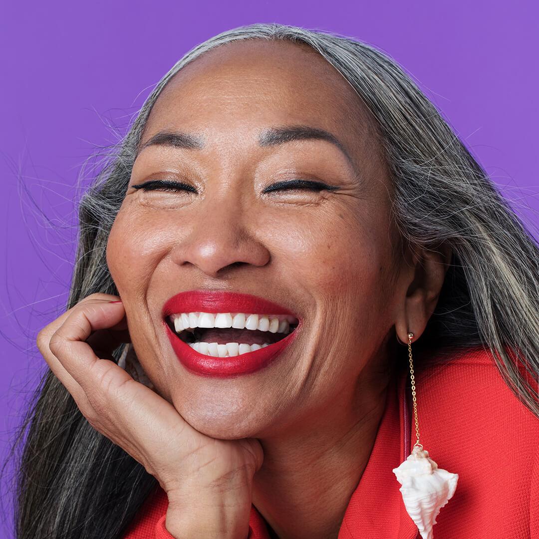 A closeup photo of a mature woman laughing wearing a red lipstick and sporting a red coat and conch earrings on a purple background Mobile
