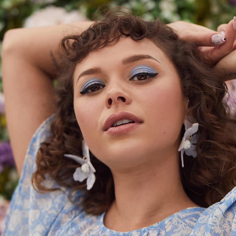 A close-up image of a model in a blue floral top rocking a light blue eyeshadow and black cat eyeliner look and wearing statement floral earrings Desktop