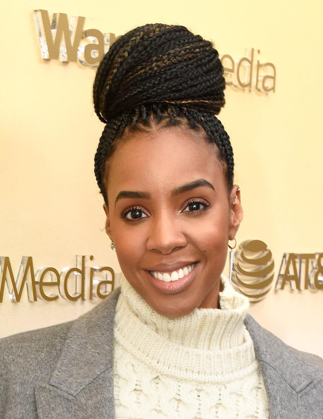 Kelly Rowland in an offwhite turtle neck sweater and grey coat rocking a braided top knot hairstyle Desktop