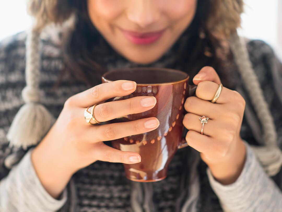 A close-up photo of a woman in a sweater, holding a brown printed mug showing her clean, clear, and polished nails adorned with golden rings in assorted shapes, including stars and triangles Desktop