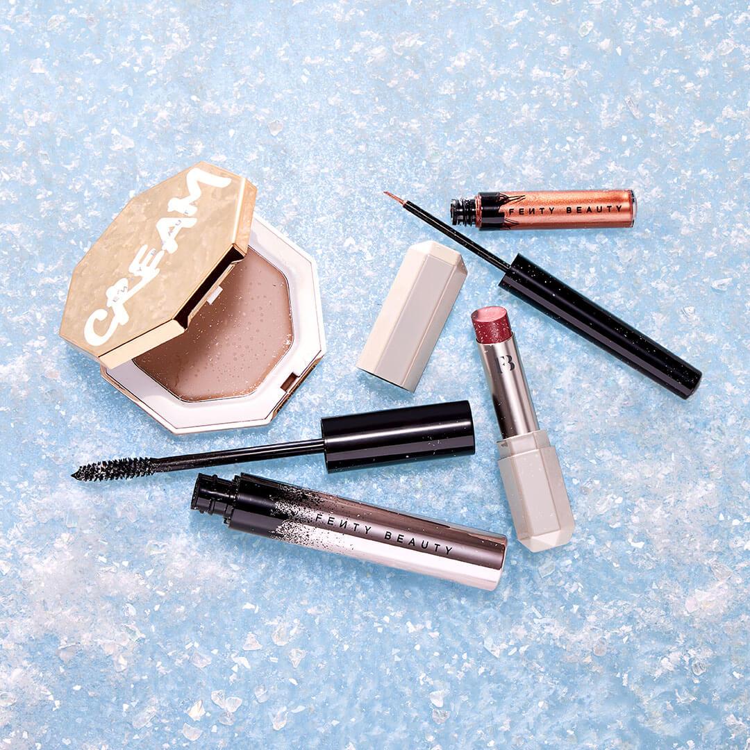 FENTY BEAUTY Holiday Bundle scattered on glittery blue surface Mobile