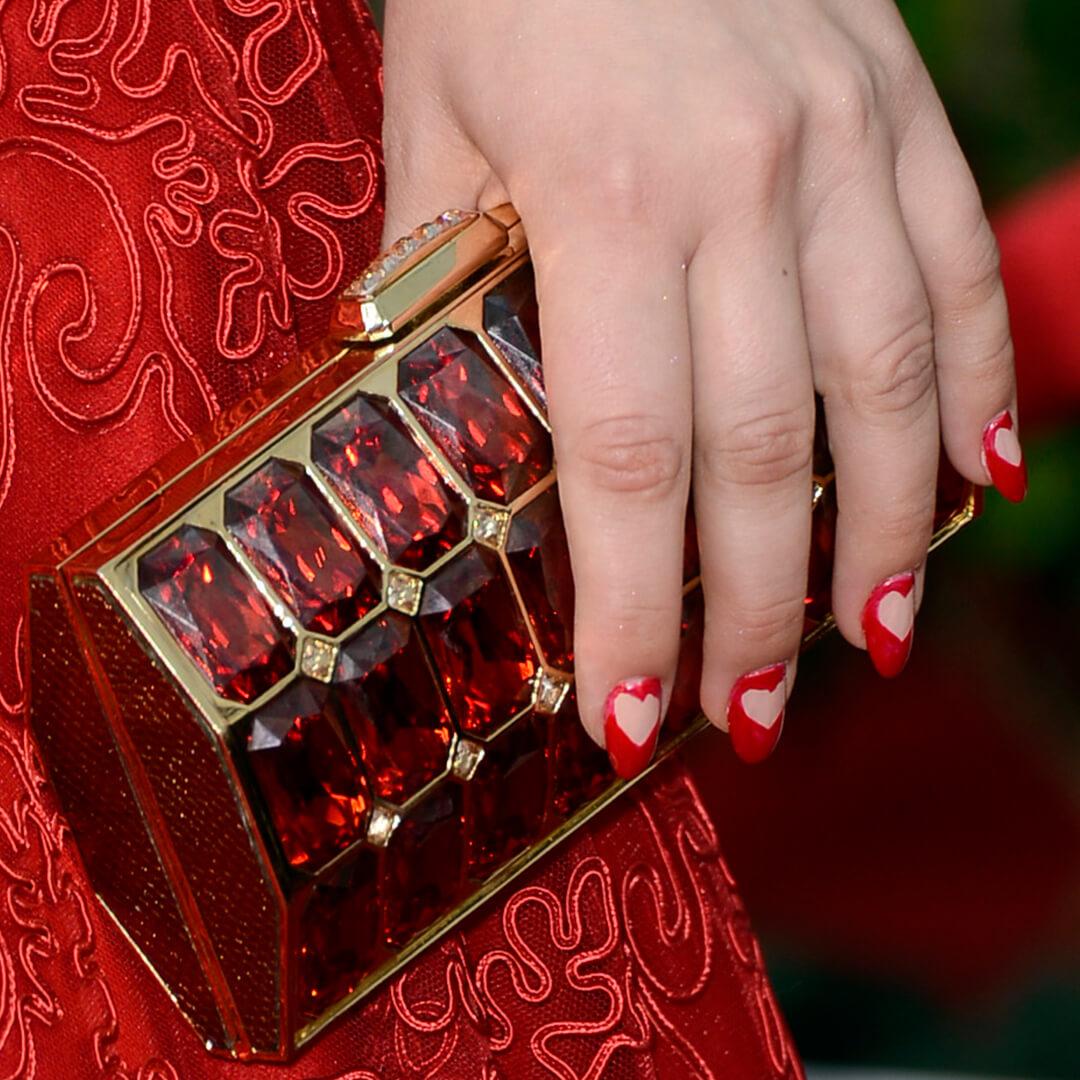 Close-up of Kaley Cuoco’s hand with negative space heart manicure holding a red-bejewelled purse Mobile