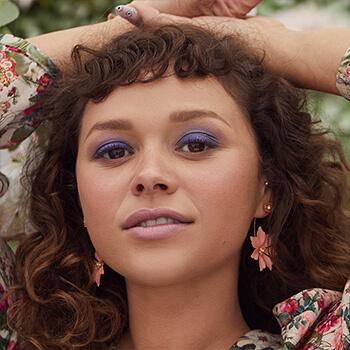 A close-up image of a curly-haired model rocking a dark blue smoky eyeshadow look and wearing floral earrings Mobile