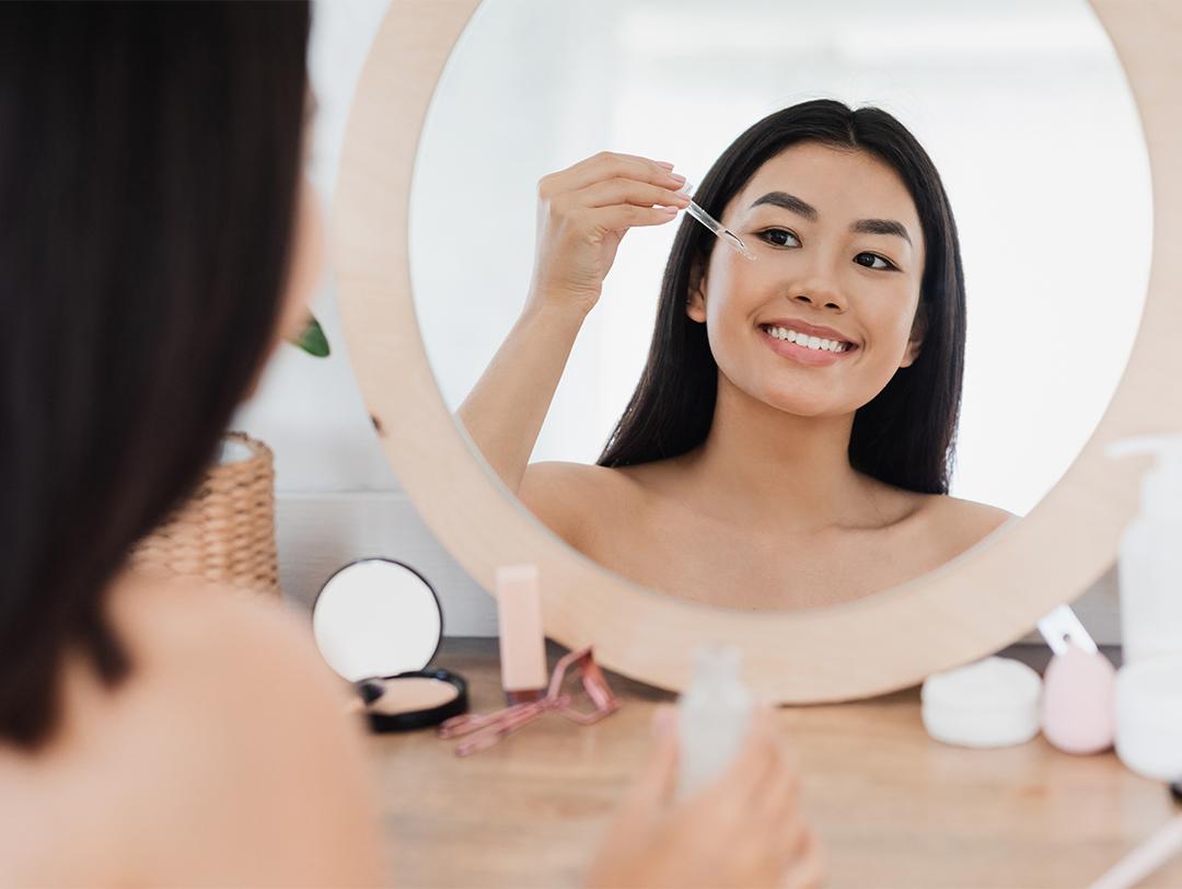 A photo of a woman facing the mirror and applying face serum Desktop