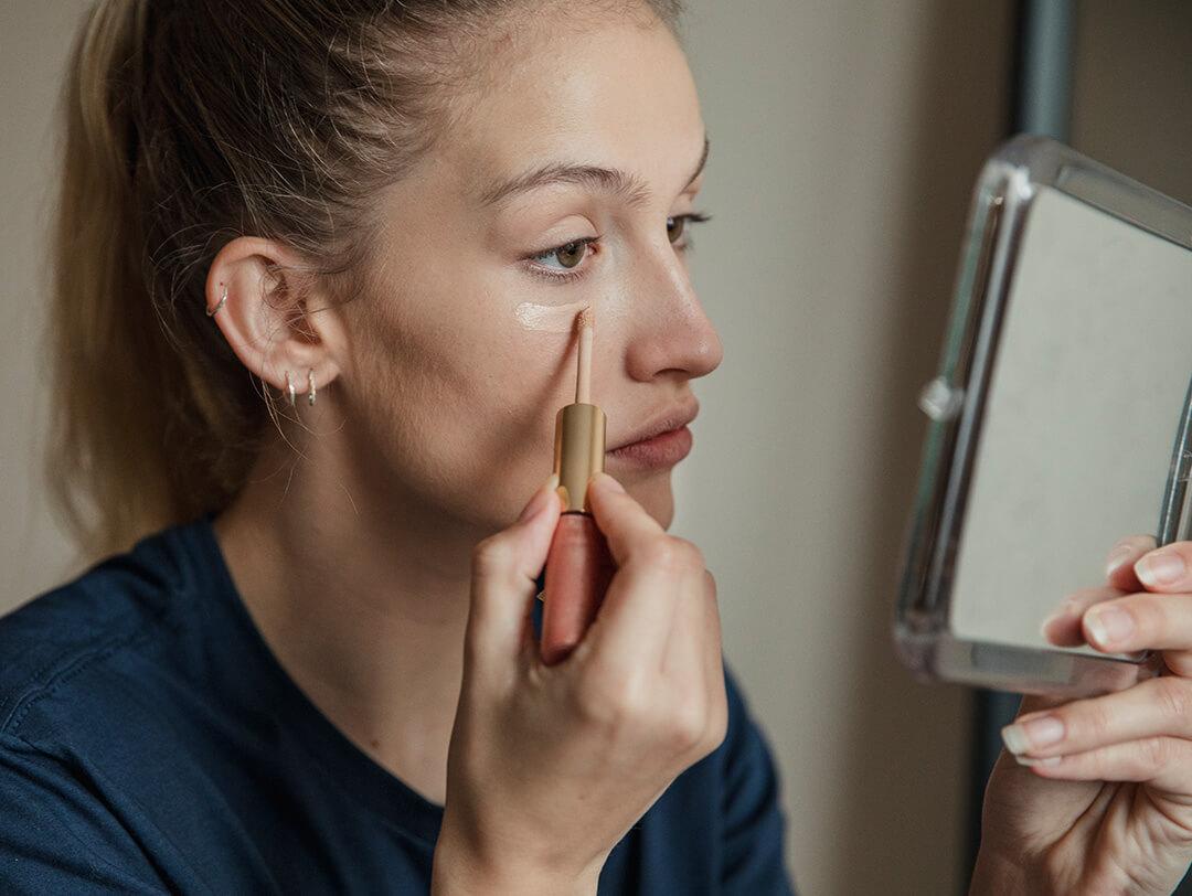 An image of a woman with her hair tied in a ponytail, gazing into a mirror while applying concealer under her eye Desktop