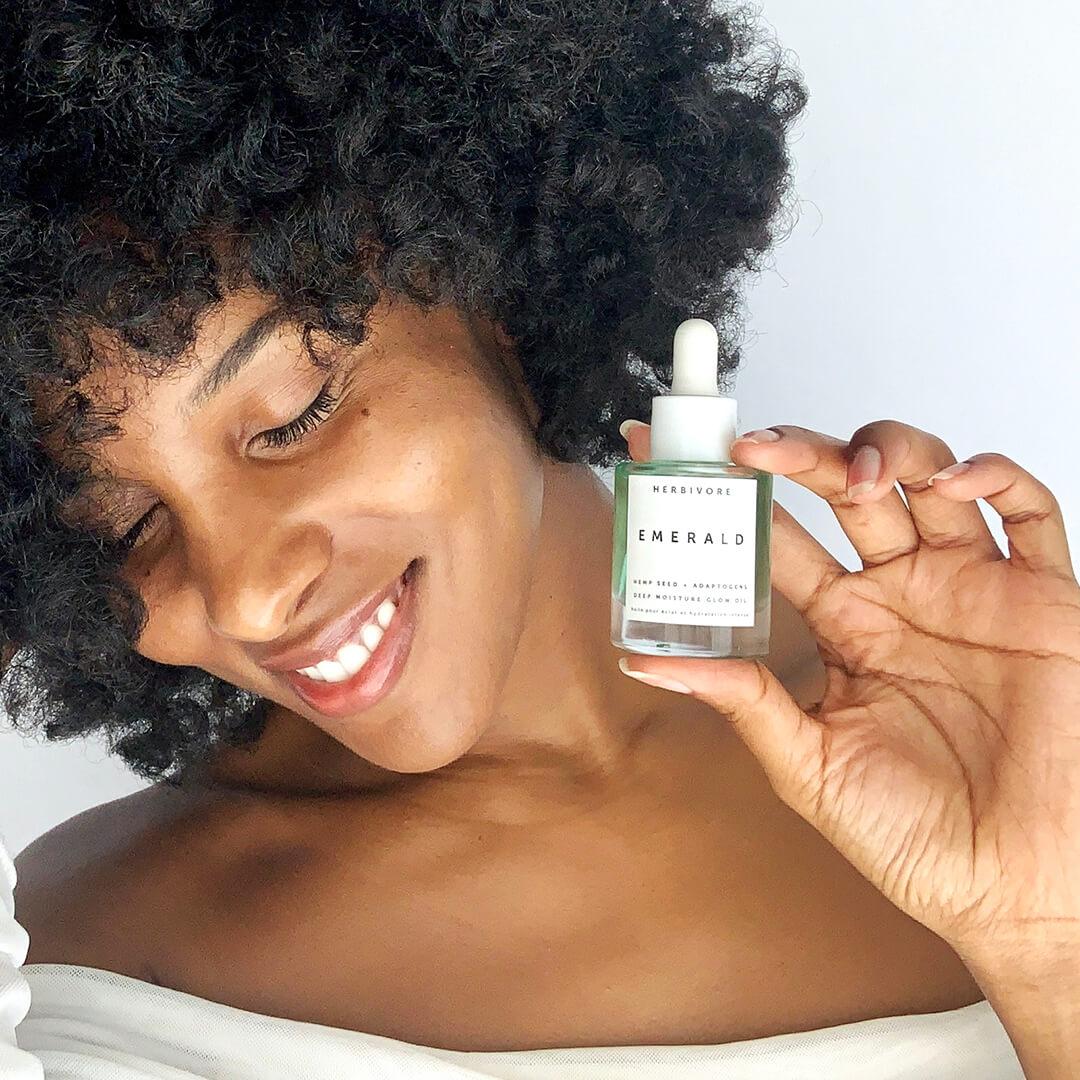 Close-up of a smiling black woman holding up the HERBIVORE Emerald Deep Moisture Glow Oil while looking down Desktop