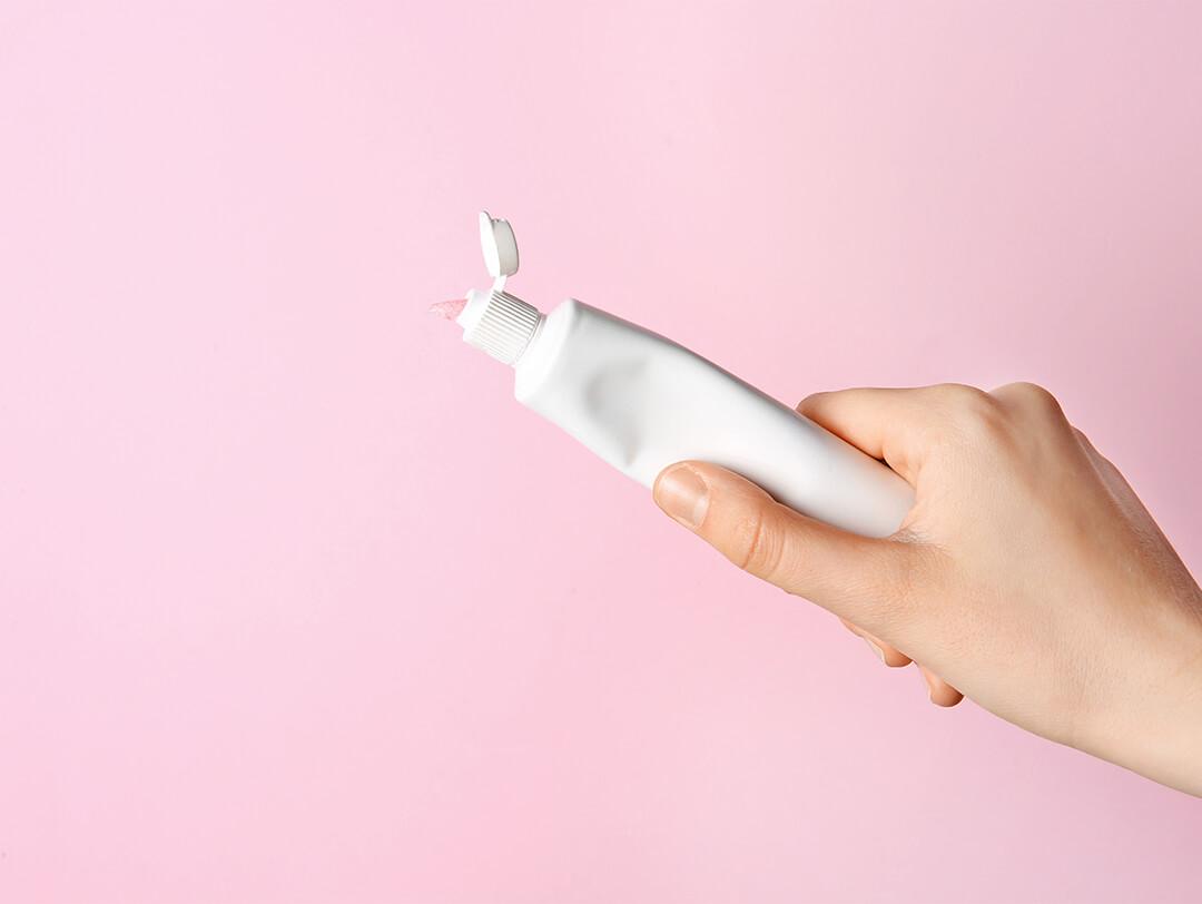 Woman's hand holding a white tube of toothpaste against pink background Desktop