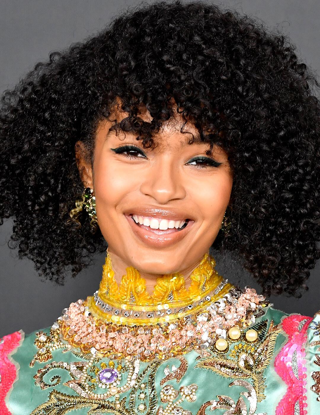 A photo of Yara Shahidi wearing a bright smile in her natural black curly hair with a curl specific haircut dressed in green full of pearls and beads Mobile