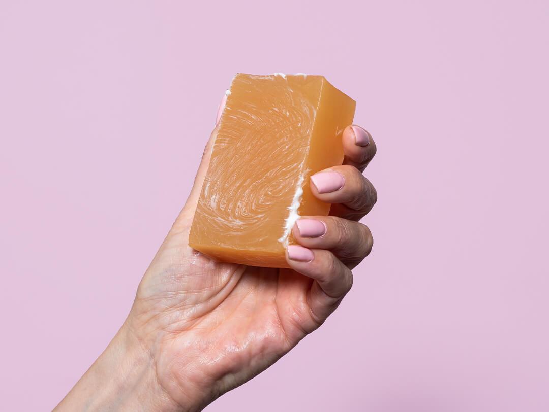 Close-up of a hand with pastel pink nail polish holding a shampoo bar against lavender background Desktop