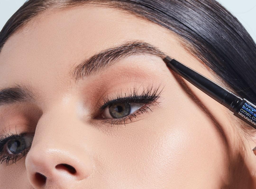 A close-up photo of a woman using a browpencil to fill in her eyebrows while wearing black eyeliner Desktop