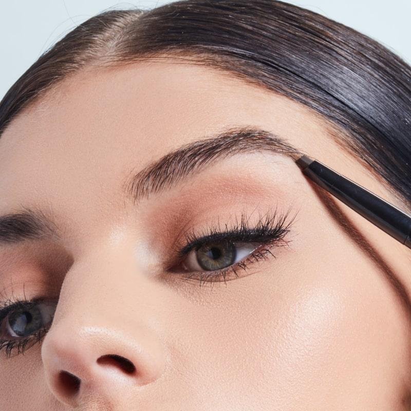 A close-up photo of a woman using a browpencil to fill in her eyebrows while wearing black eyeliner Mobile