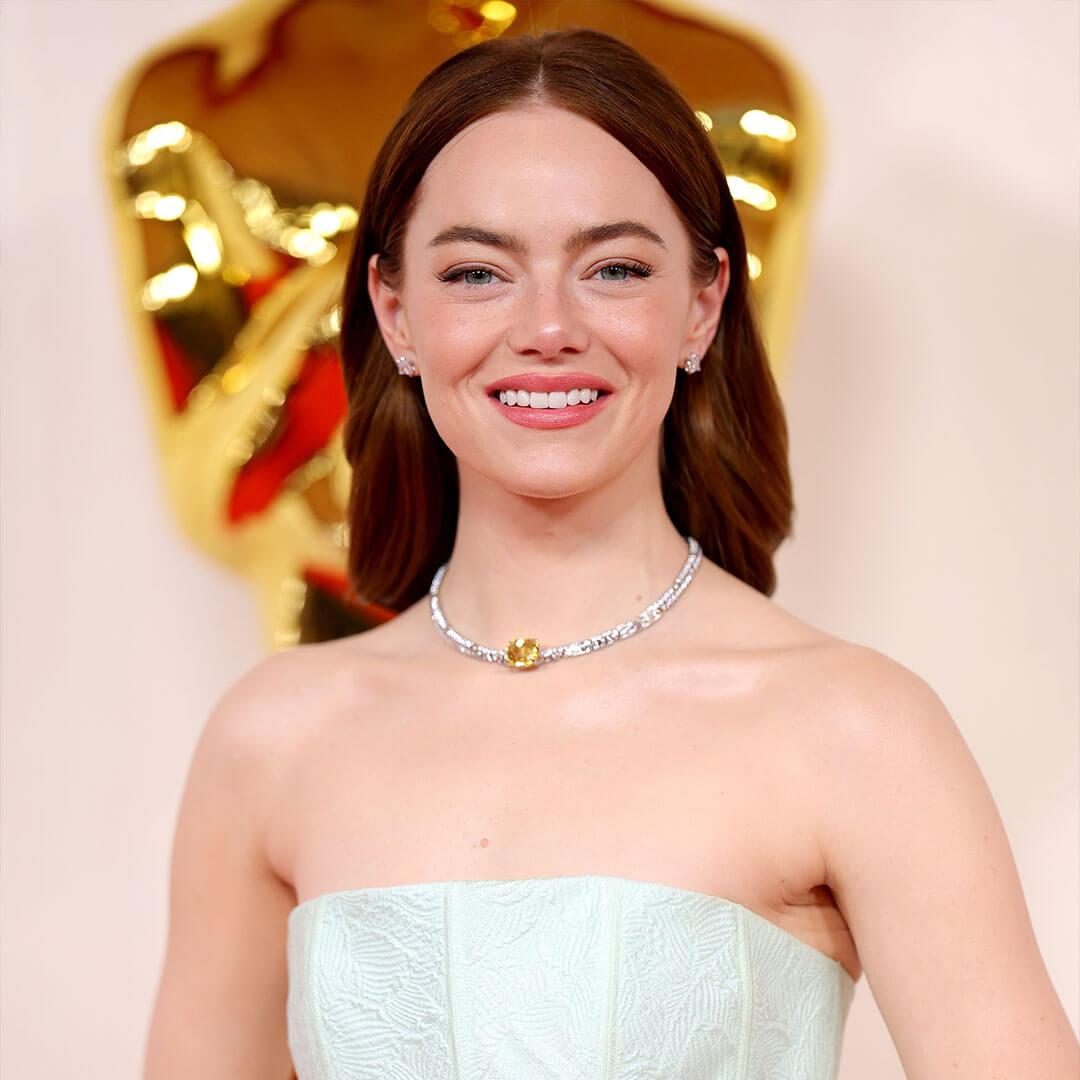 A photo of Emma Stone grinning at the camera while donning a pale pink monochromatic makeup and a white off-the-shoulder sheath gown Mobile
