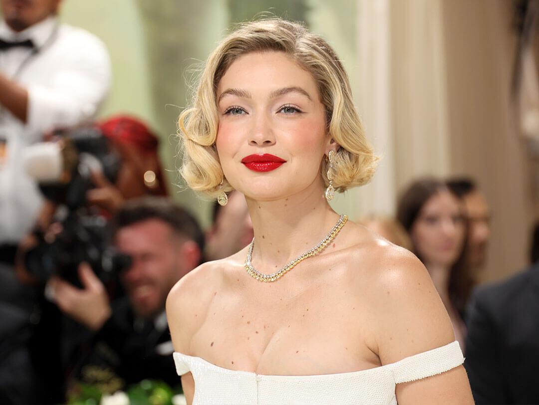 A photo of Gigi Hadid showing off her short blond hair, bright red lipstick, white off-the-shoulder dress, and diamond gold necklace Desktop