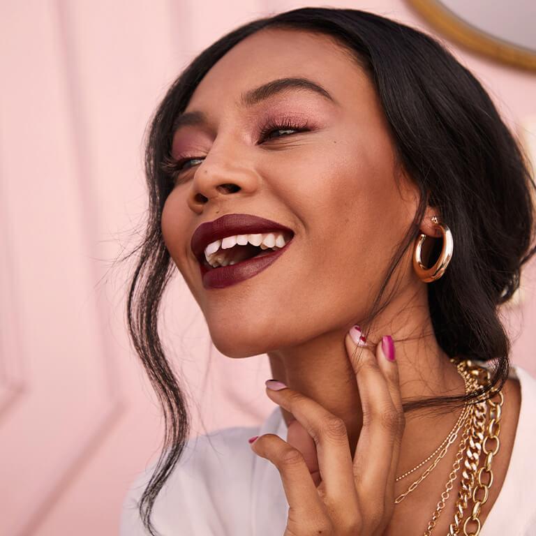 An image of a model wearing gold jewelry, rose gold eyeshadow, and dark berry lipstick smiling big and holding her neck with two fingers Desktop
