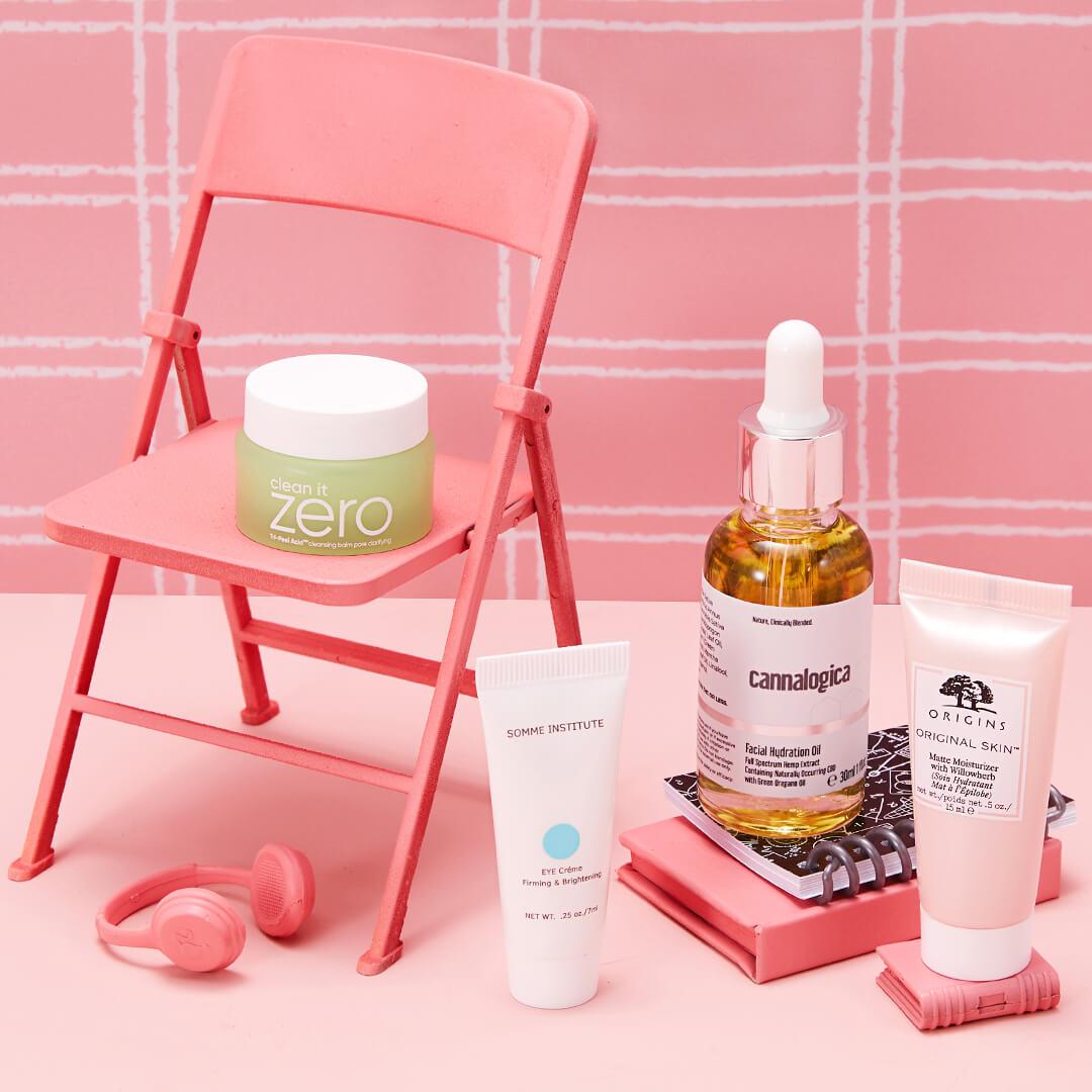 Flat-lay image of skincare products from the September 2021 IPSY Glam Bag and Glam Bag Plus on miniature pink chair, notebooks, headphones, against pink tile background Desktop