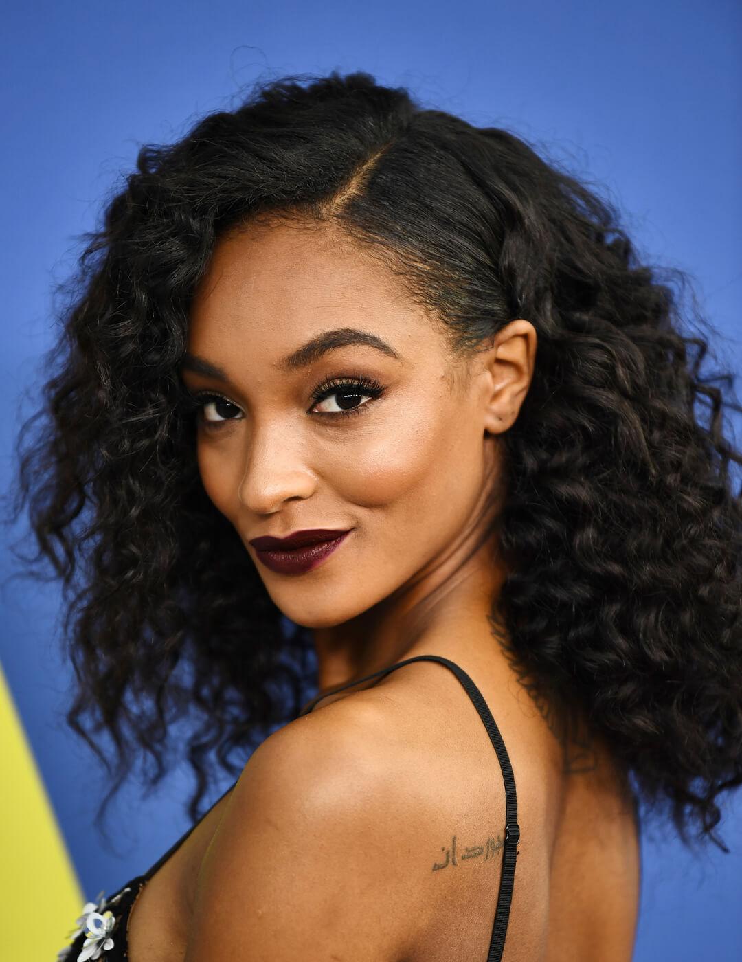 Jourdan Dunn in a black spaghetti strap dress rocking a parted, curly hairstyle while smirking Mobile