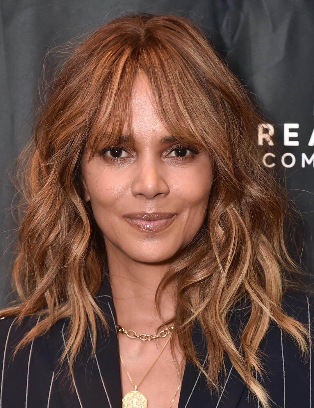 A photo of Halle Berry with dark blonde highlight extentions wearing a black coat with white stripes and gold chain necklace Mobile