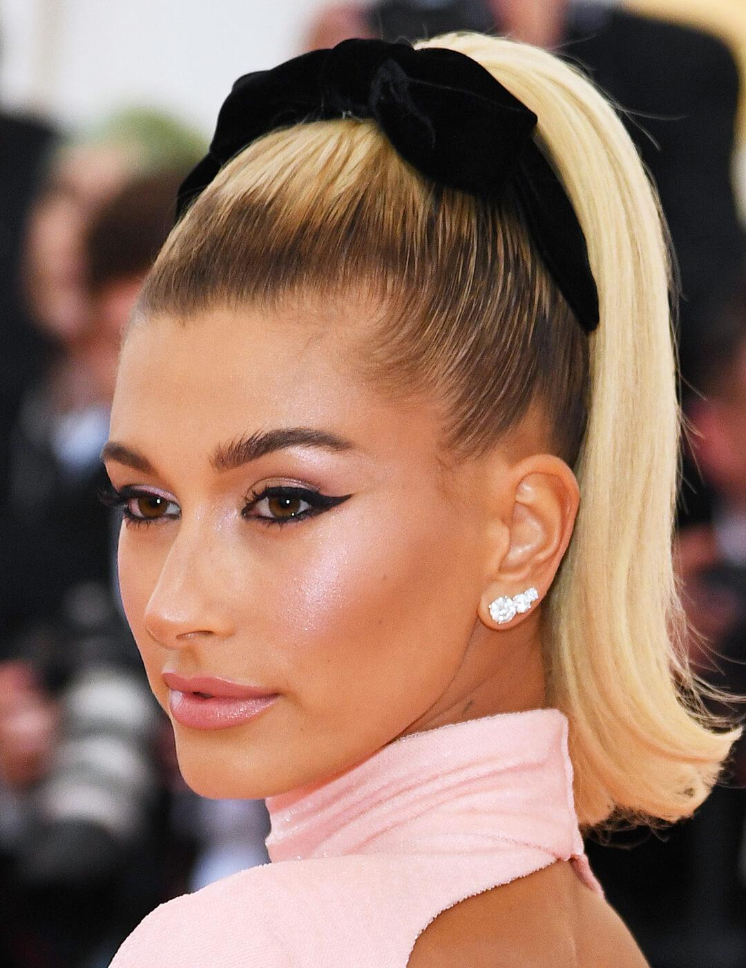 Hailey Bieber rocking a sleek, high ponytail hairstyle, bold winged eyeliner makeup look, diamond earrings, and pink dress Mobile