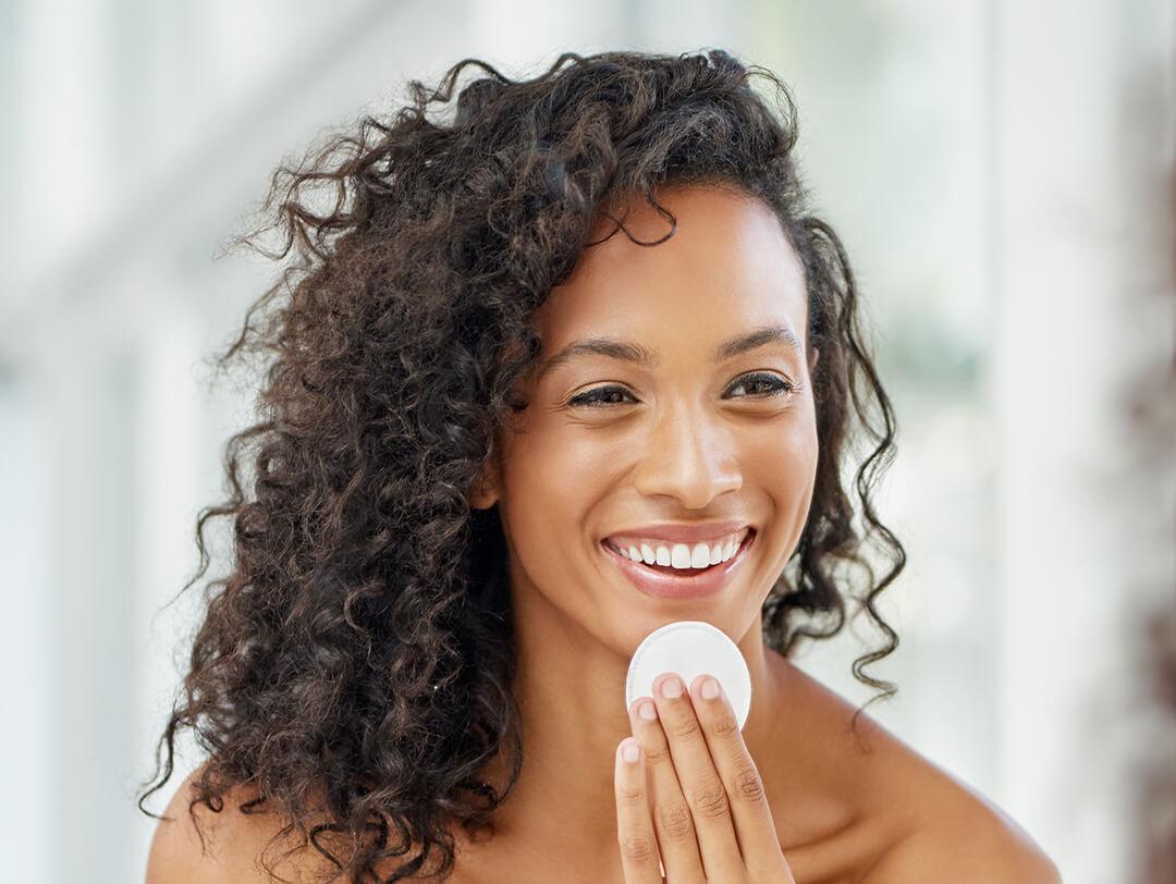 An image of a curly-haired woman smiling while applying a skincare product on her bare face Desktop