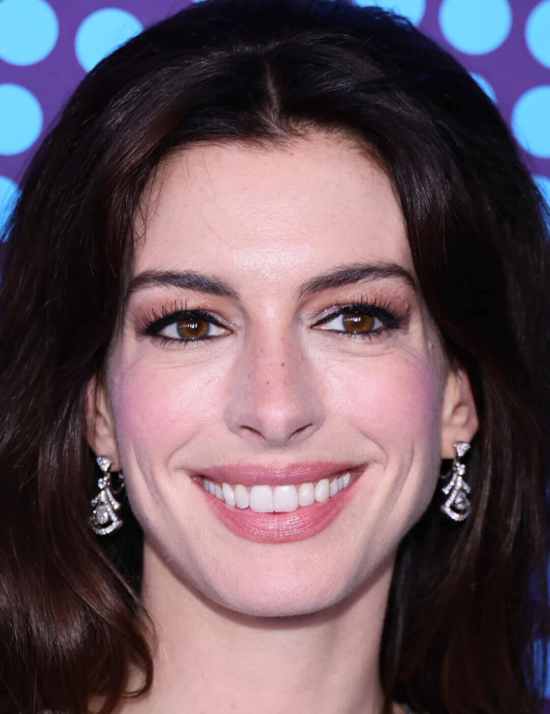 A close-up photo of Anne Hathaway with dark brown straight hair, and subtle makeup that accentuates her downturned eyes with black eyeliner, elegantly dressed in a pearl-beaded gown and wearing dangling earrings Mobile