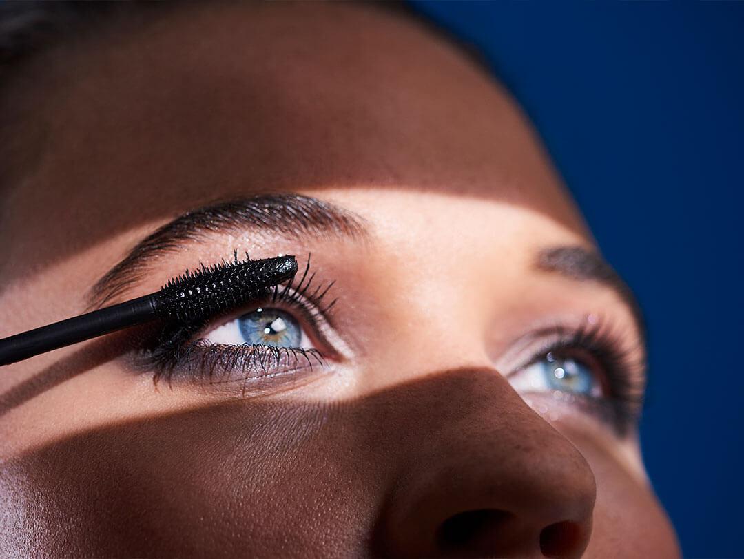 A close-up shot of a woman with blue eyes, with dramatic shadow across part of her face, as she applies black mascara to her upper lashes, brushing them upward to create a lifted, curled effect Desktop