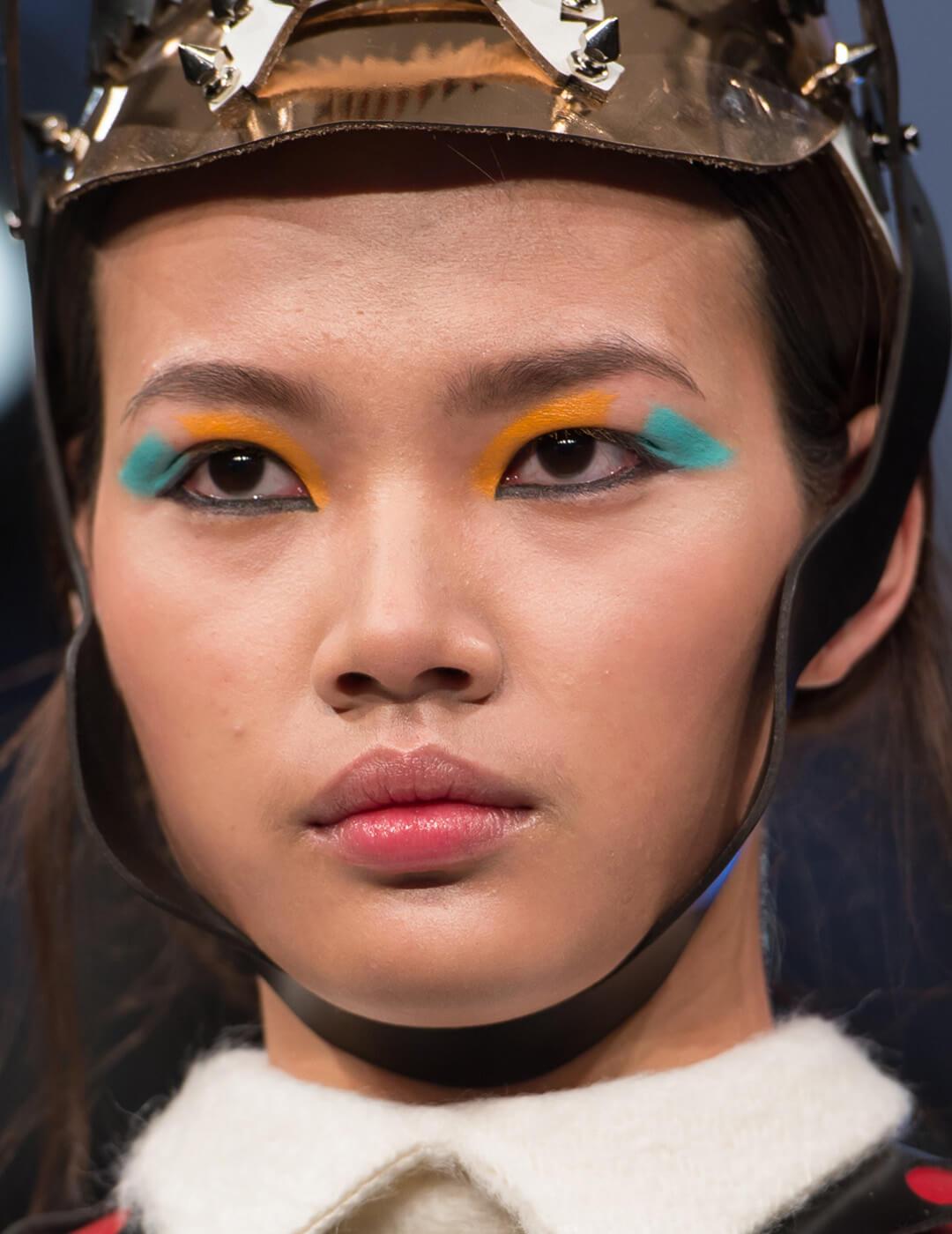 Model wearing a brown leather head piece and blue and orange eye makeup Mobile
