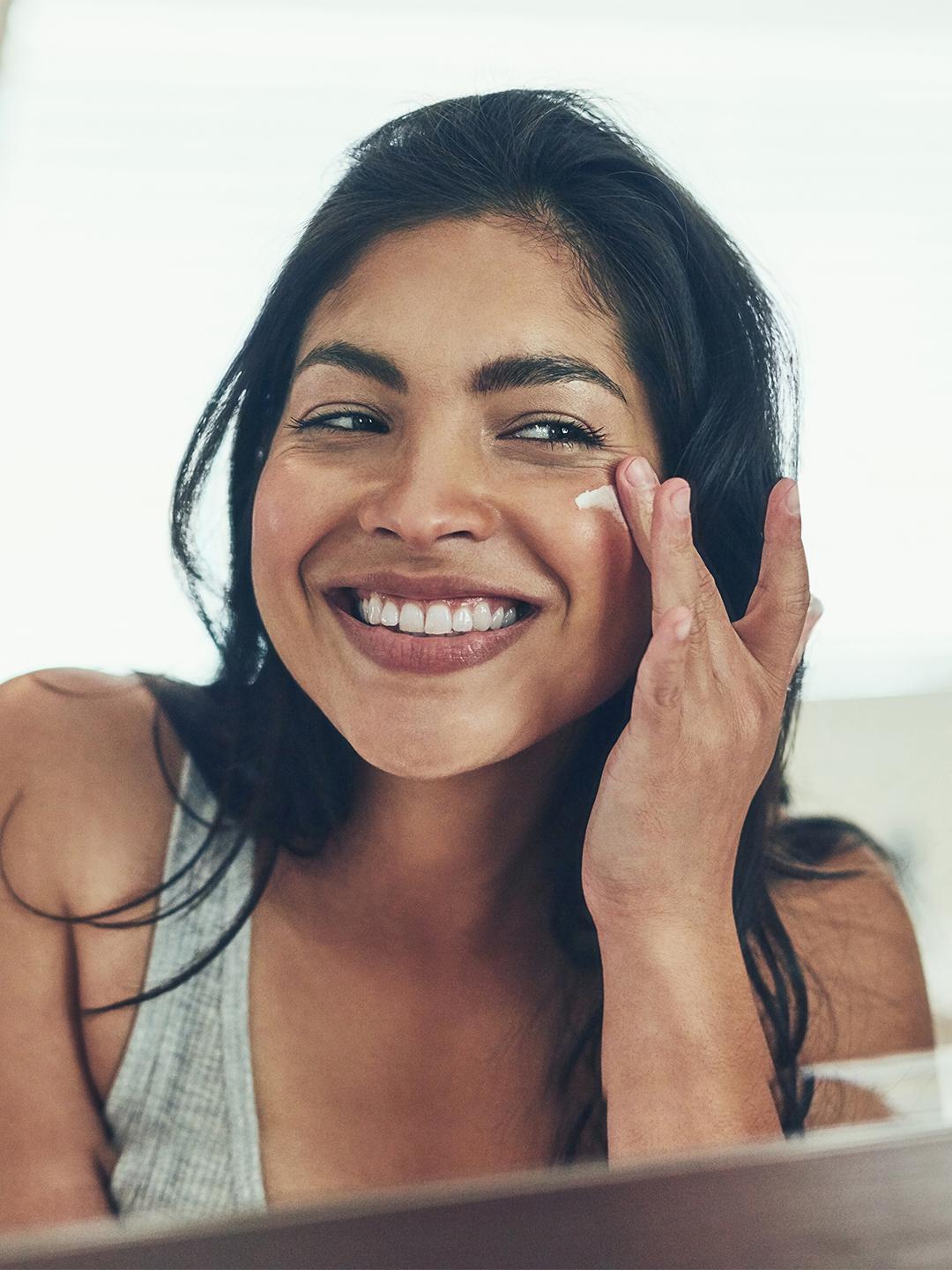 Image of a woman in a grey tank top putting on cream on her cheek Desktop