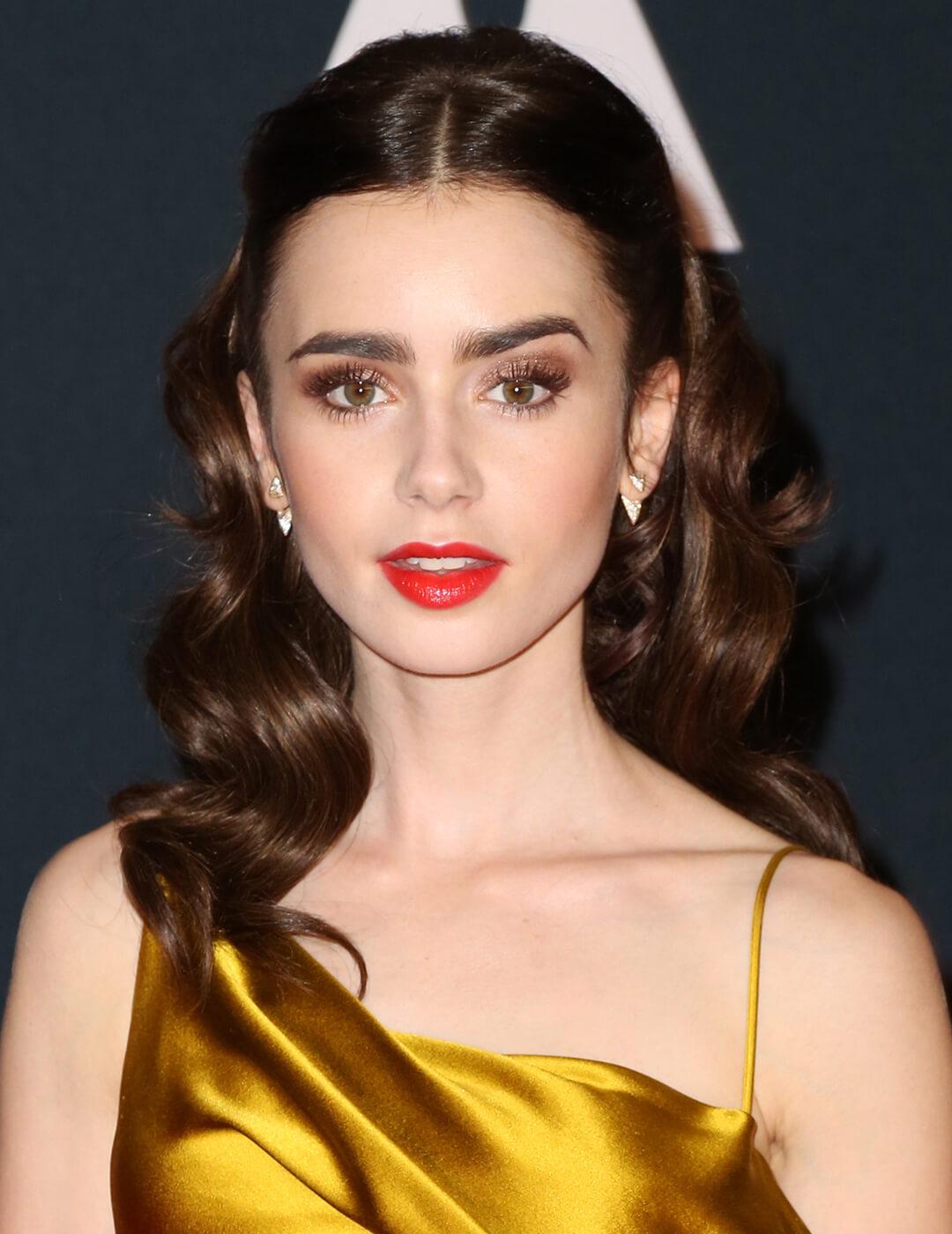 Lily Collins looking elegant in a neutral makeup look and red lips, golden satin dress, and wavy hair Mobile