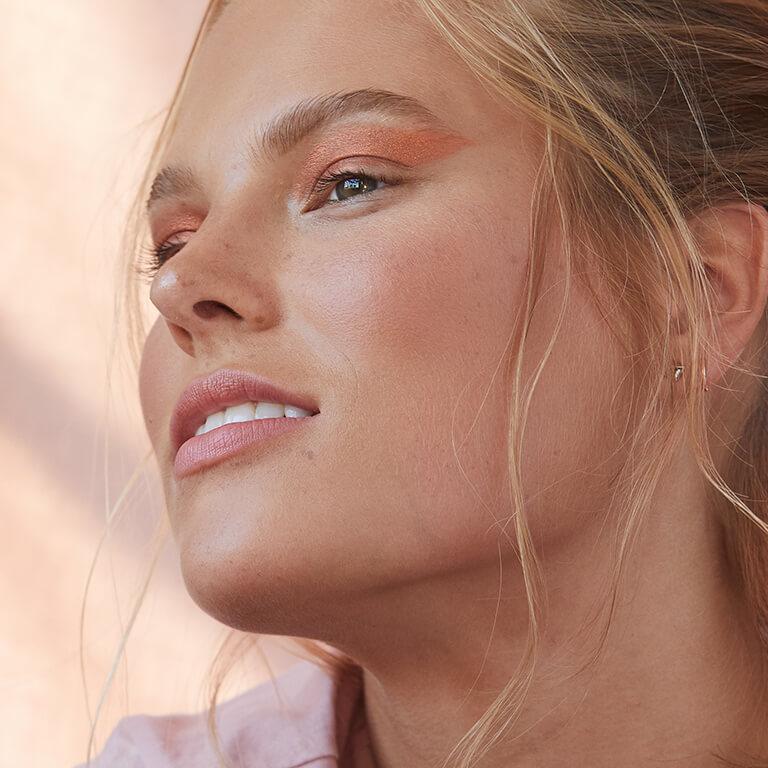 A closeup profile image of a model wearing shimmery copper eyeshadow paired with glossy pink lipstick Mobile