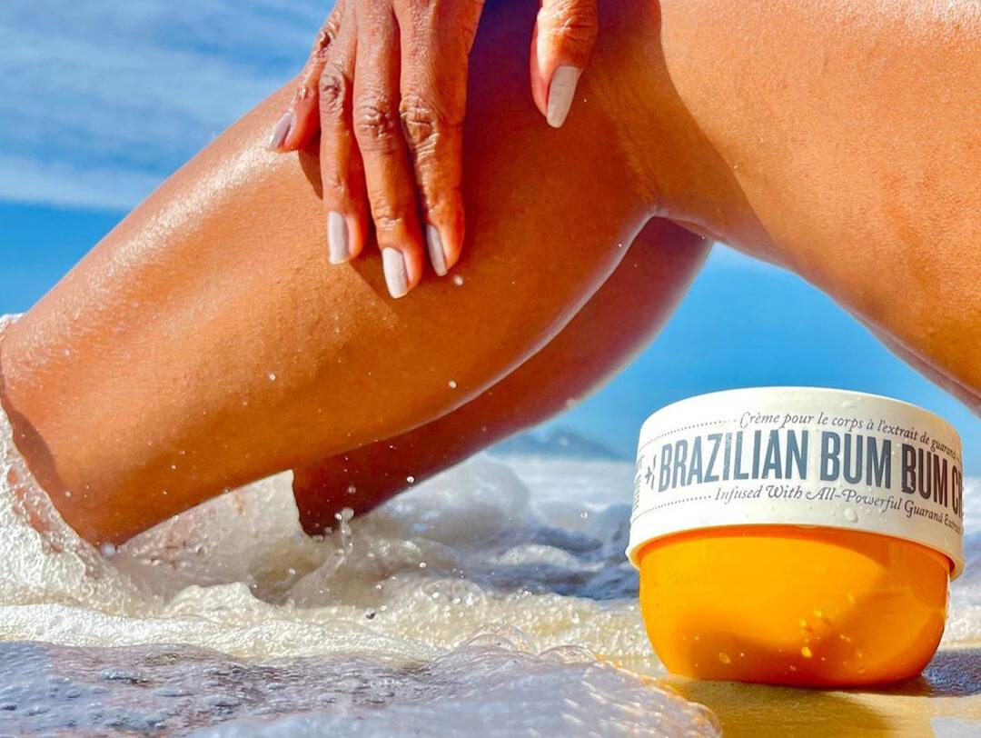 Close-up of SOL DE JANEIRO Brazilian Bum Bum Cream on the sand by the water beside a woman's legs Desktop