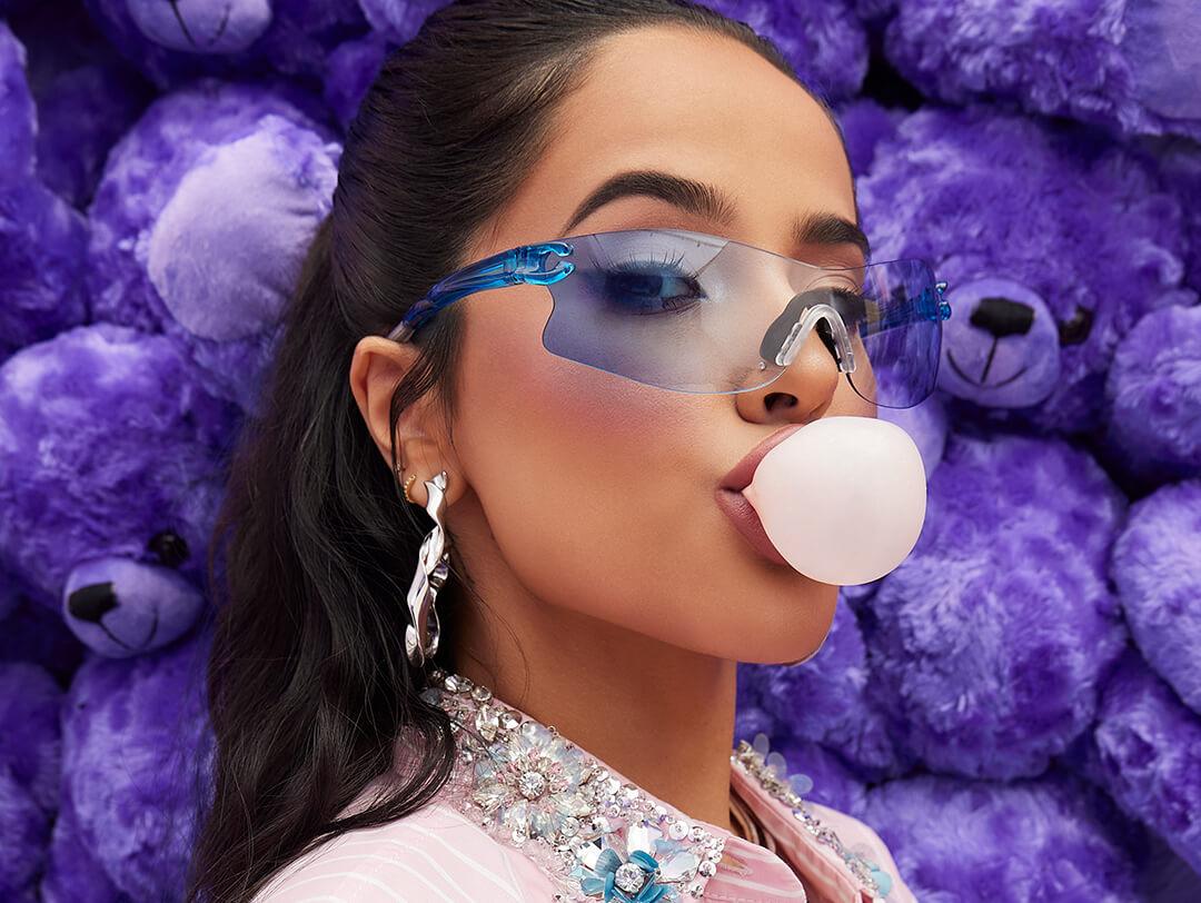 Close-up of Becky G wearing blue sunglasses and blowing a bubble gum against a wall of blue teddy bears Desktop