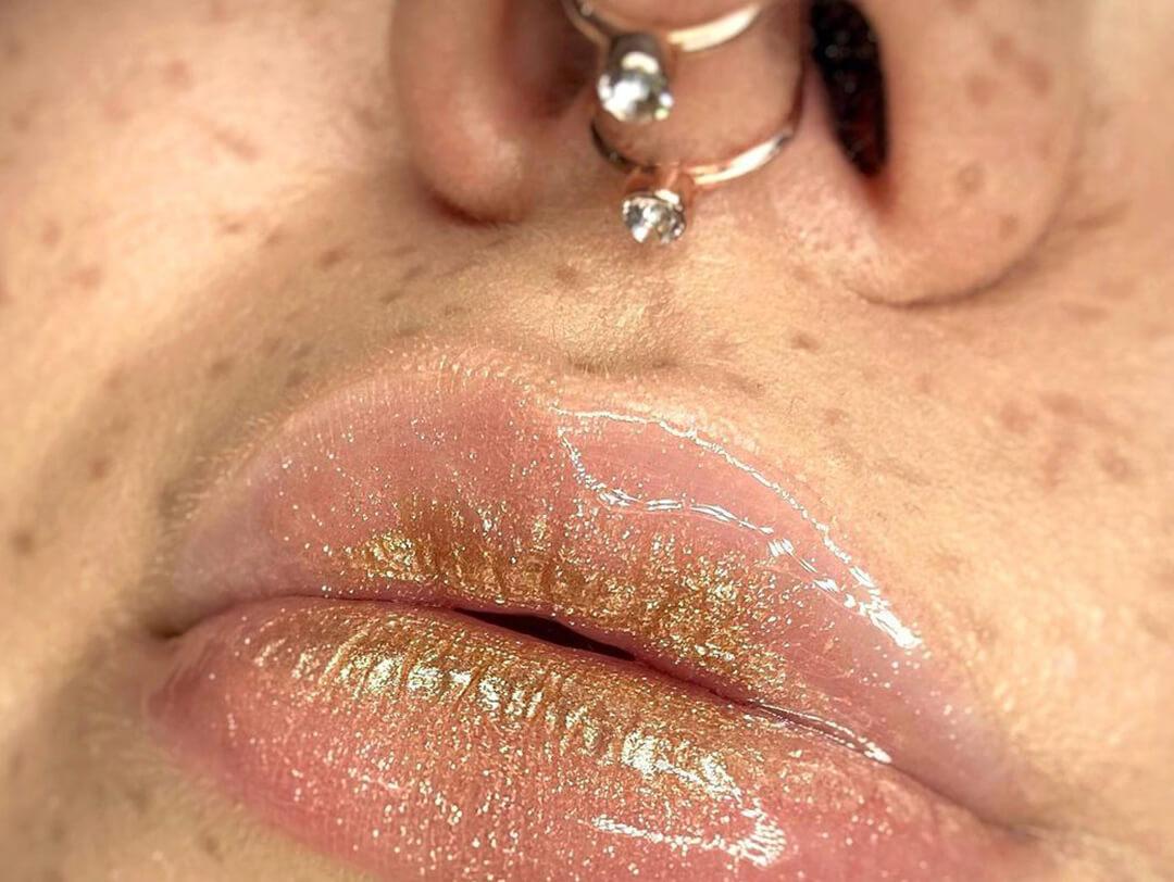 A close-up image of a woman's lips coated with golden clear lip gloss showing a diamond nose piercing Desktop