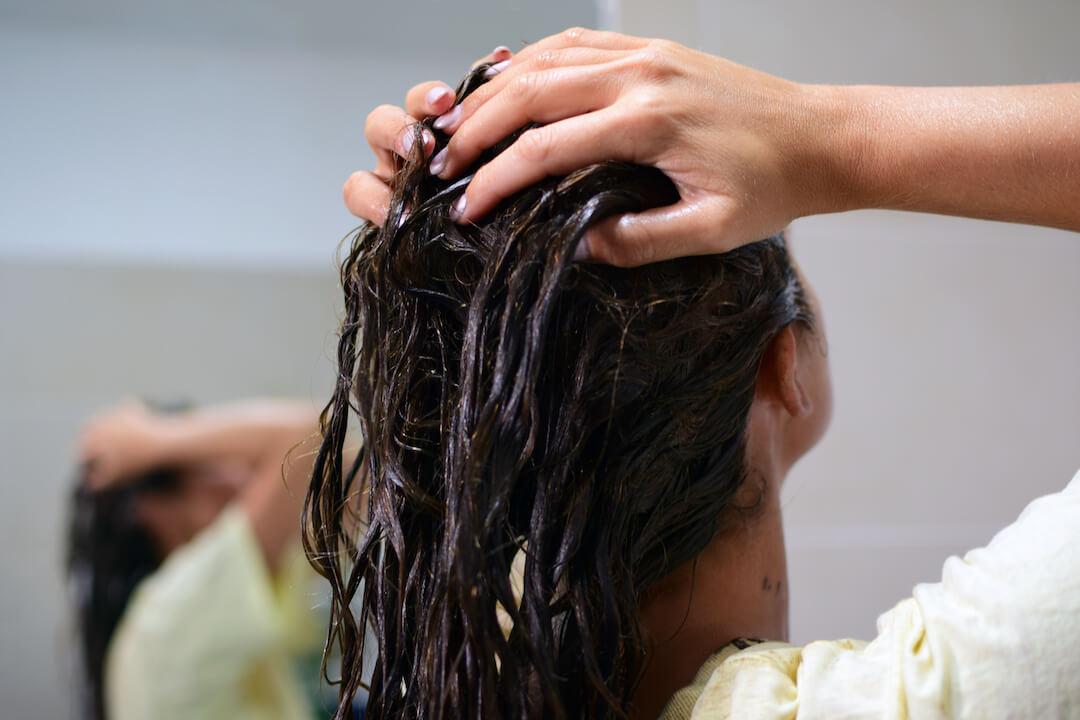 An image of a woman with black hair facing away while applying hair dye, showcasing her purple gel nails Desktop