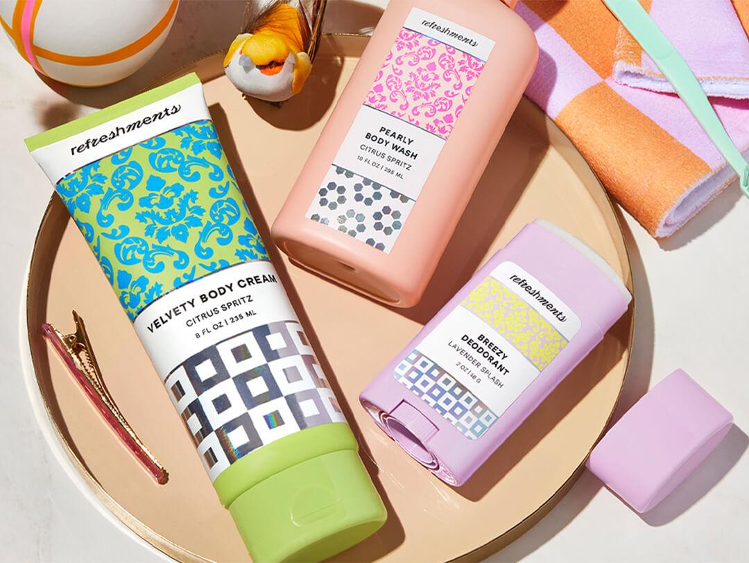 Flat-lay image of REFRESHMENTS Velvety Body Cream in Citrus Spritz, Pearly Body Wash in Citrus Spritz, and Breezy Deodorant in Lavender Splash on cream-colored plate Desktop