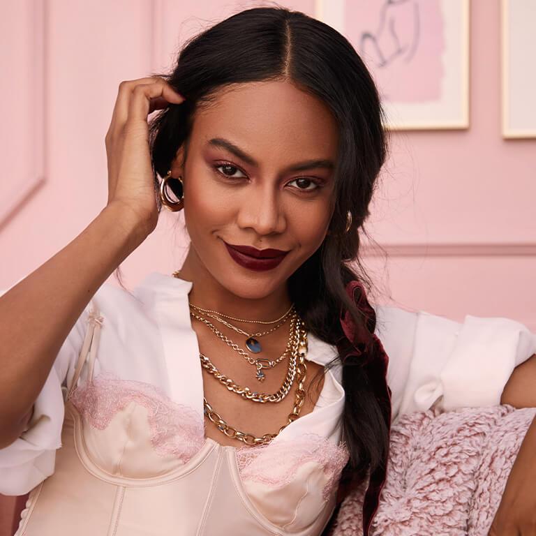 An image of a smirking model with her French braid to her side wearing gold jewelry and pulling one side of her bangs behind her ear Desktop