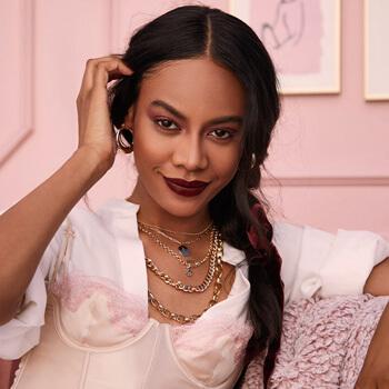 An image of a smirking model with her French braid to her side wearing gold jewelry and pulling one side of her bangs behind her ear Mobile