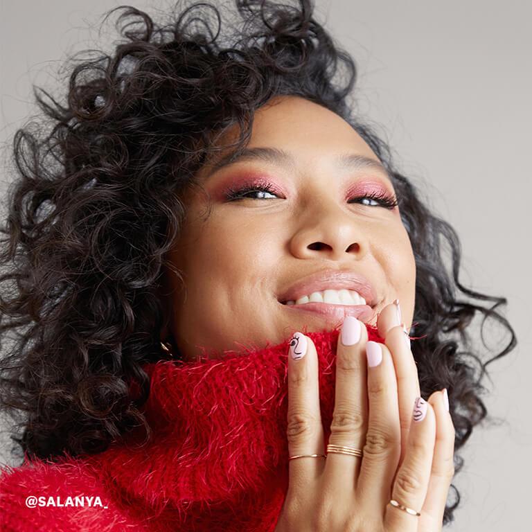 An image of a model with curly hair and hands together wearing red, shimmery eye shadow and a red, wool turtleneck sweater Desktop