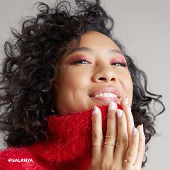 An image of a model with curly hair and hands together wearing red, shimmery eye shadow and a red, wool turtleneck sweater Mobile